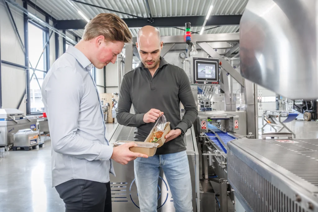 Twee Sismatec-medewerkers controleren verpakte trayseal voedingsproducten bij een industriële verpakkingslijn.