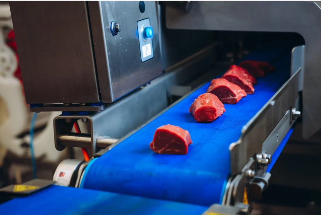 Stukken rauw vlees op een blauwe transportband richting een industriële vleessnijmachine.